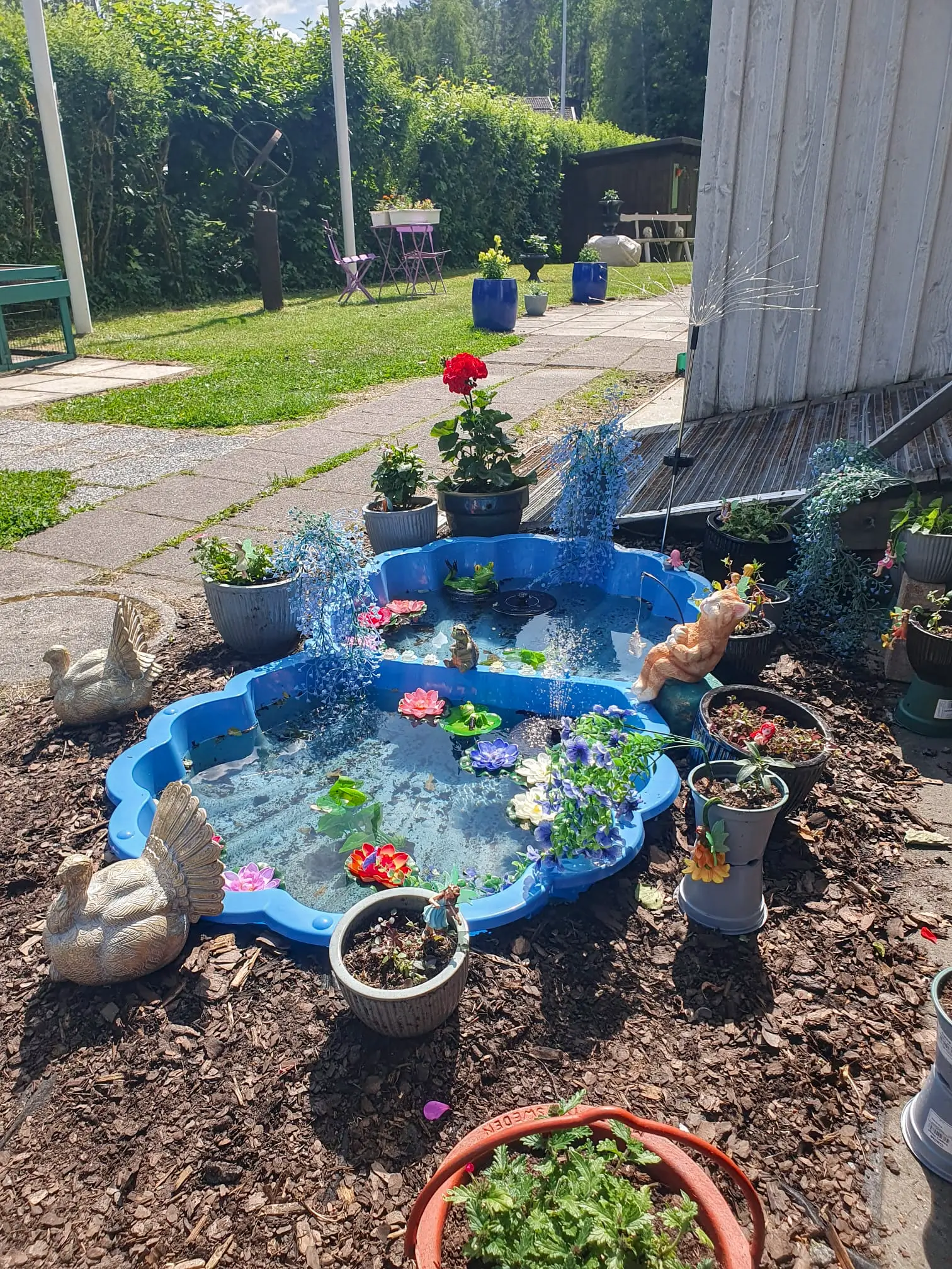 A charming garden display at Motel E4 featuring a small blue decorative pond with artificial water lilies, surrounded by swan figurines, bark mulch, and various potted flowers.