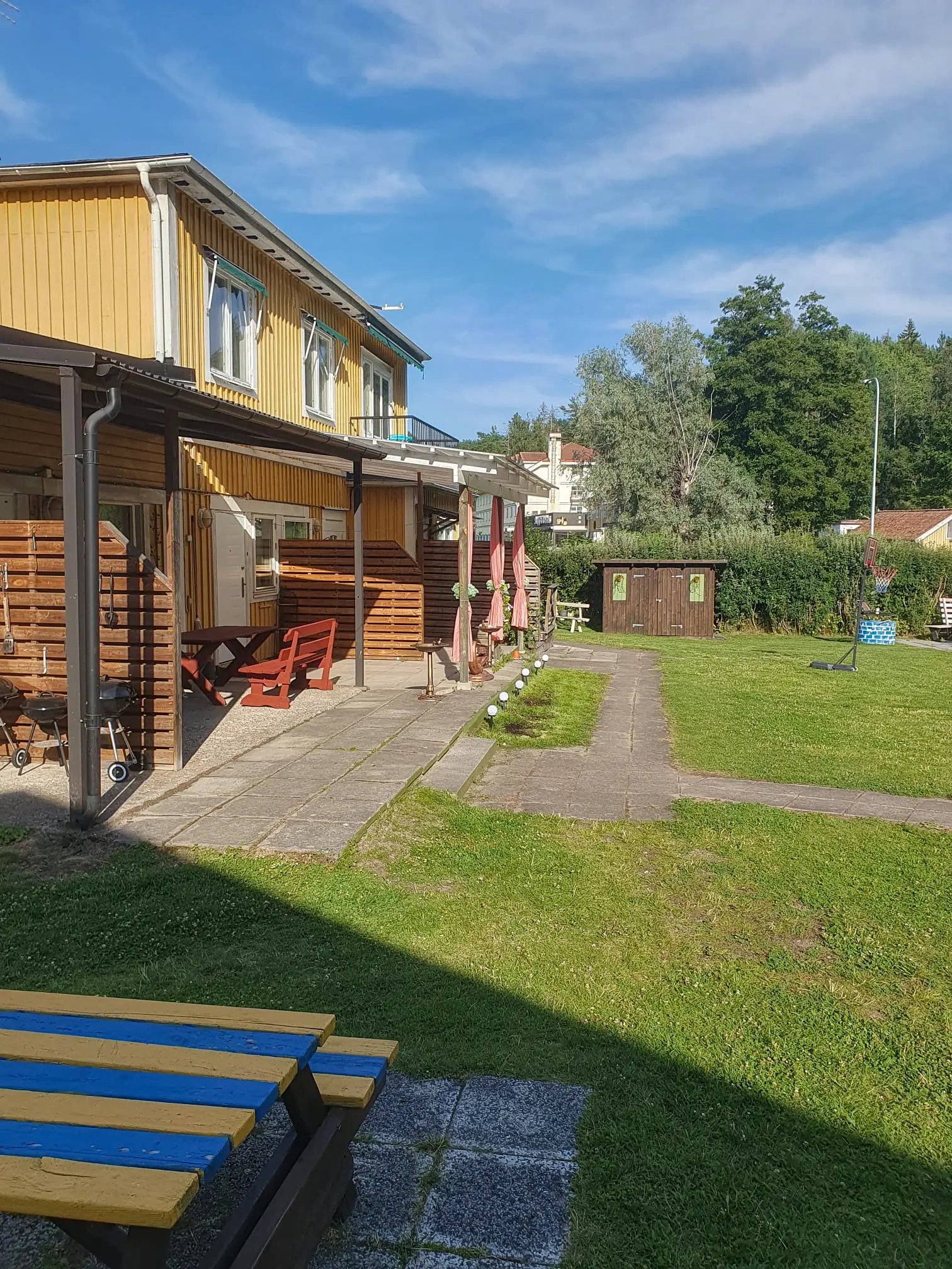 Outdoor view of the yellow guest rooms at Motel E4, featuring private patios with red furniture and a spacious green lawn area with a basketball hoop.