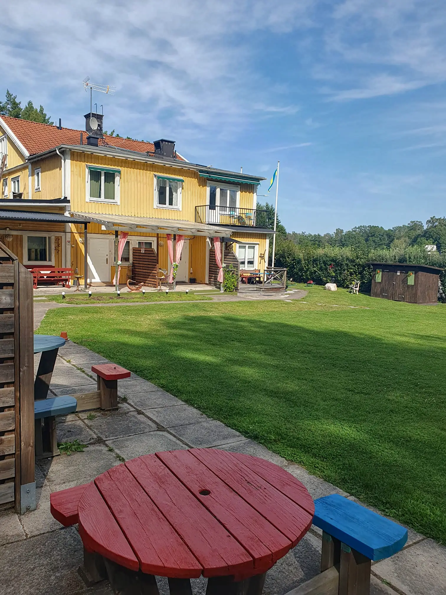 A scenic outdoor area at Motel E4, featuring a red picnic table in the foreground, a large green lawn, and the yellow motel building under a clear blue sky.
