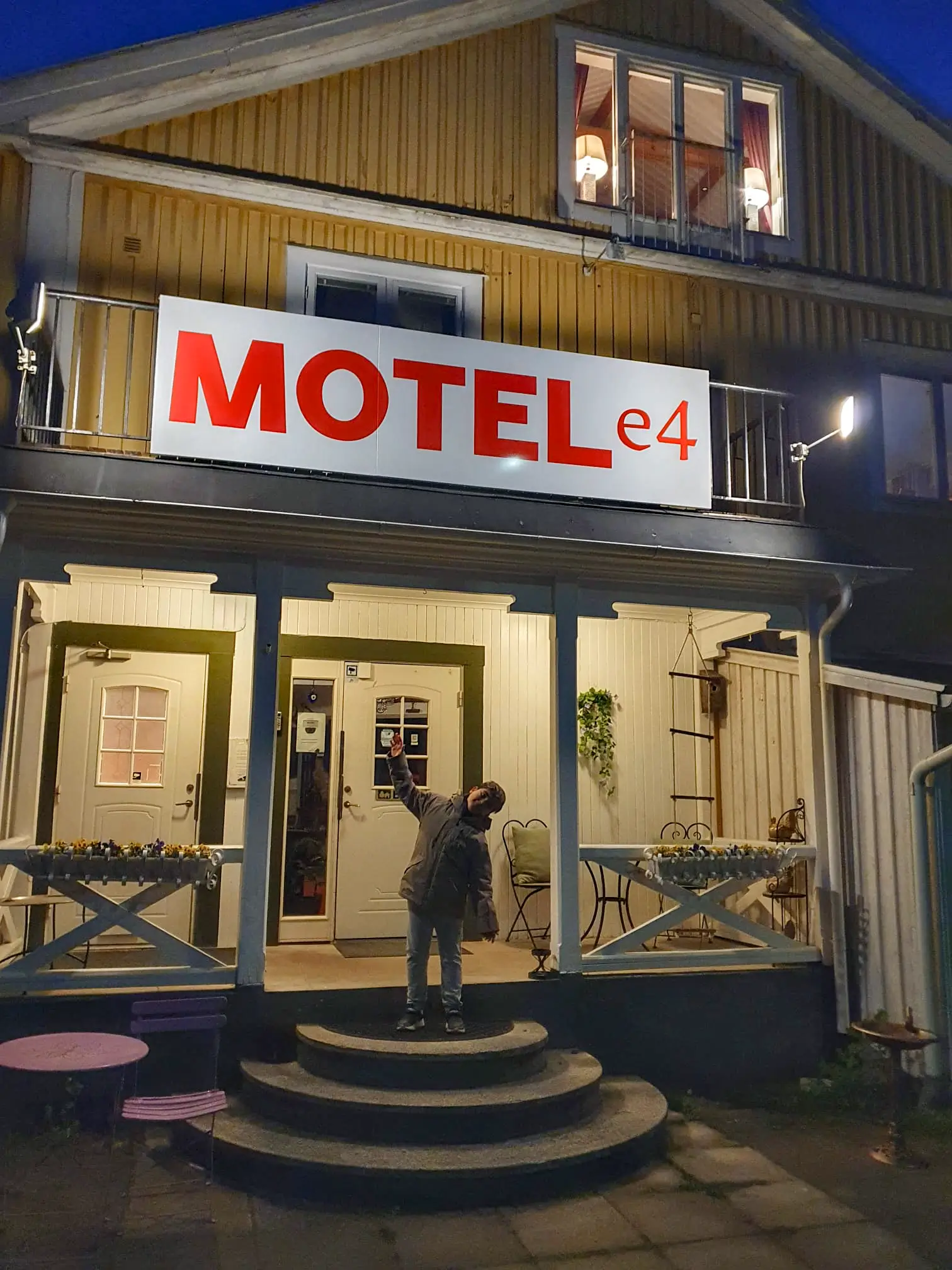 An evening view of the illuminated Motel E4 entrance, featuring its large red and white sign and a child playfully posing on the stone steps of the front porch.