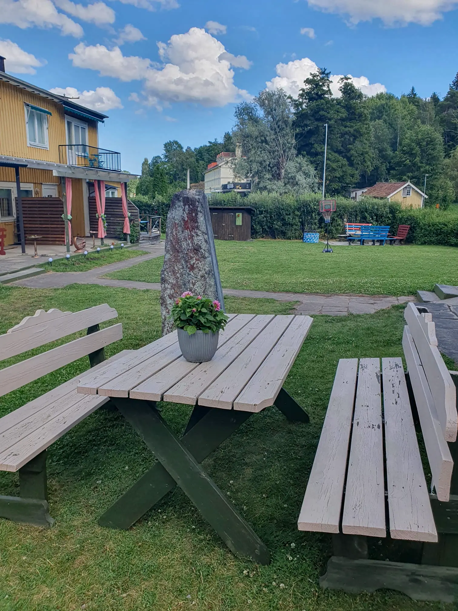 A peaceful outdoor seating area at Motel E4 featuring a wooden picnic table, a large stone monument, and a spacious green lawn under a sunny, cloud-filled sky.