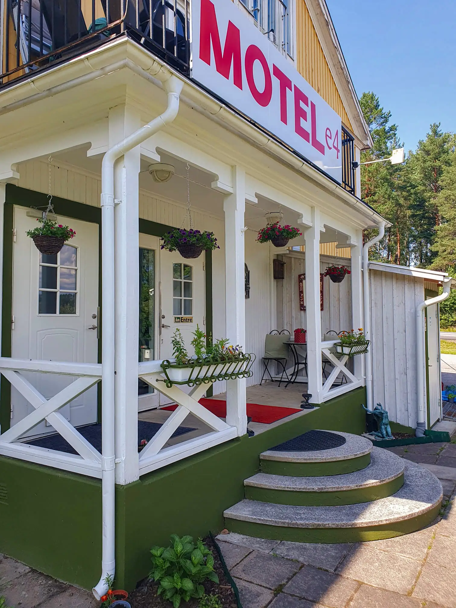 The inviting main entrance of Motel e4, featuring a white wooden porch decorated with hanging flowers, stone steps, and a cozy seating area.