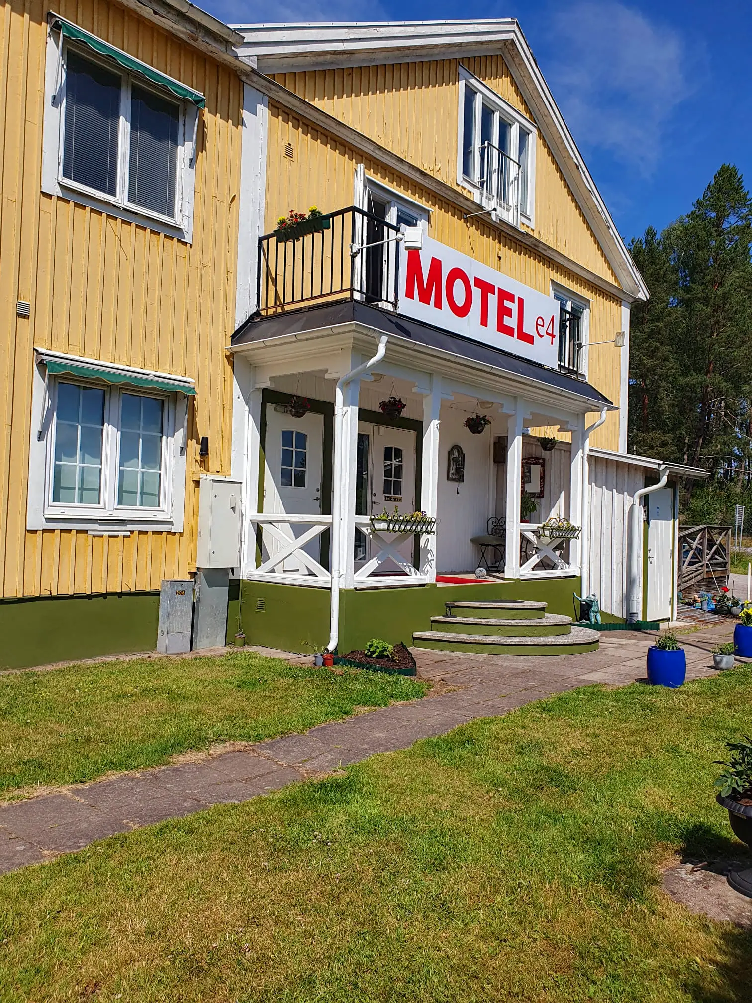 The front entrance of Motel e4, featuring its yellow wooden exterior, a white porch with steps, and the red and white "MOTEL e4" sign on a sunny day.