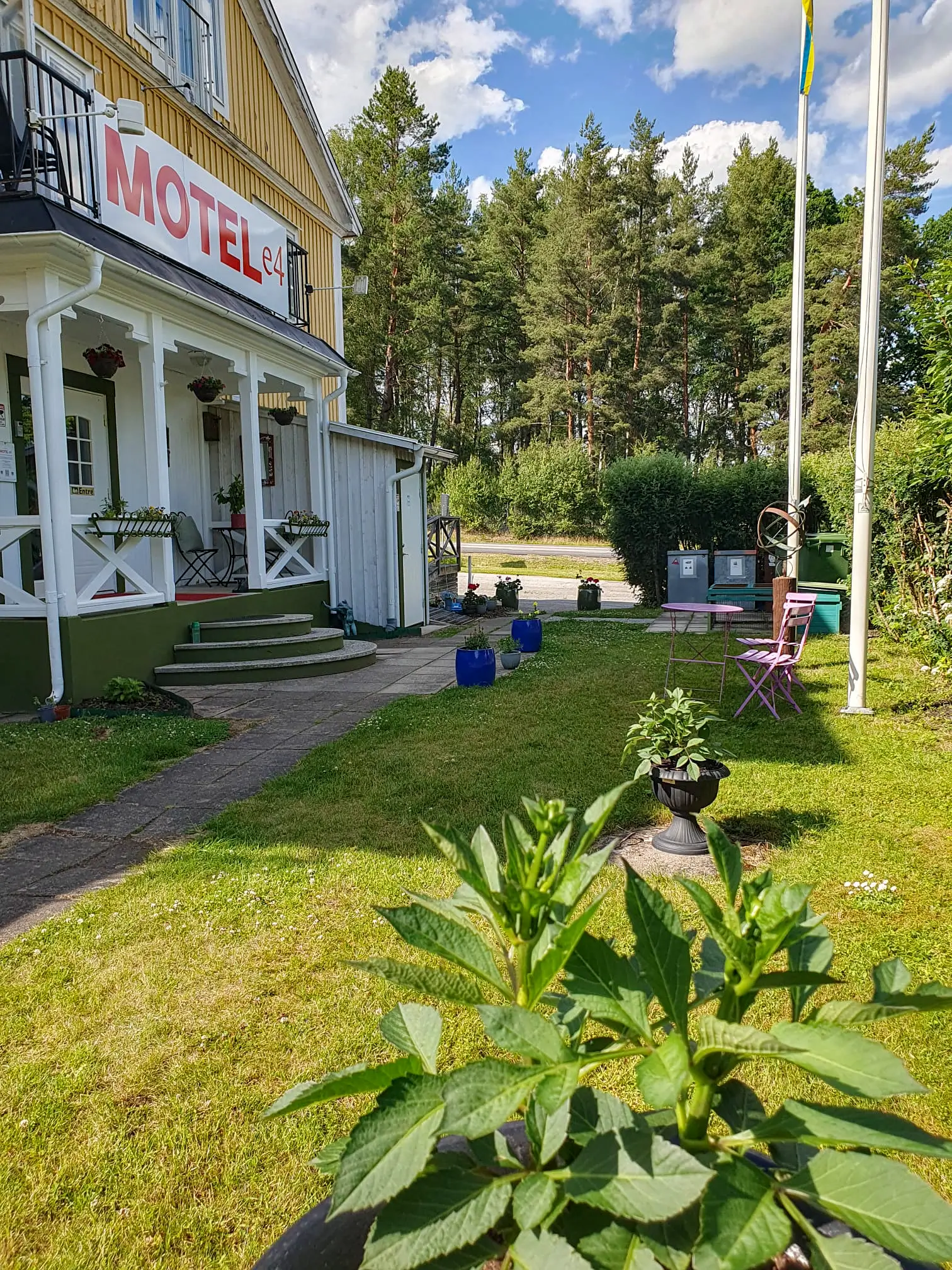 The welcoming exterior of Motel E4 in Stavsjö, featuring a bright yellow building with its red and white sign, a cozy white porch, and a sunny lawn with seating.
