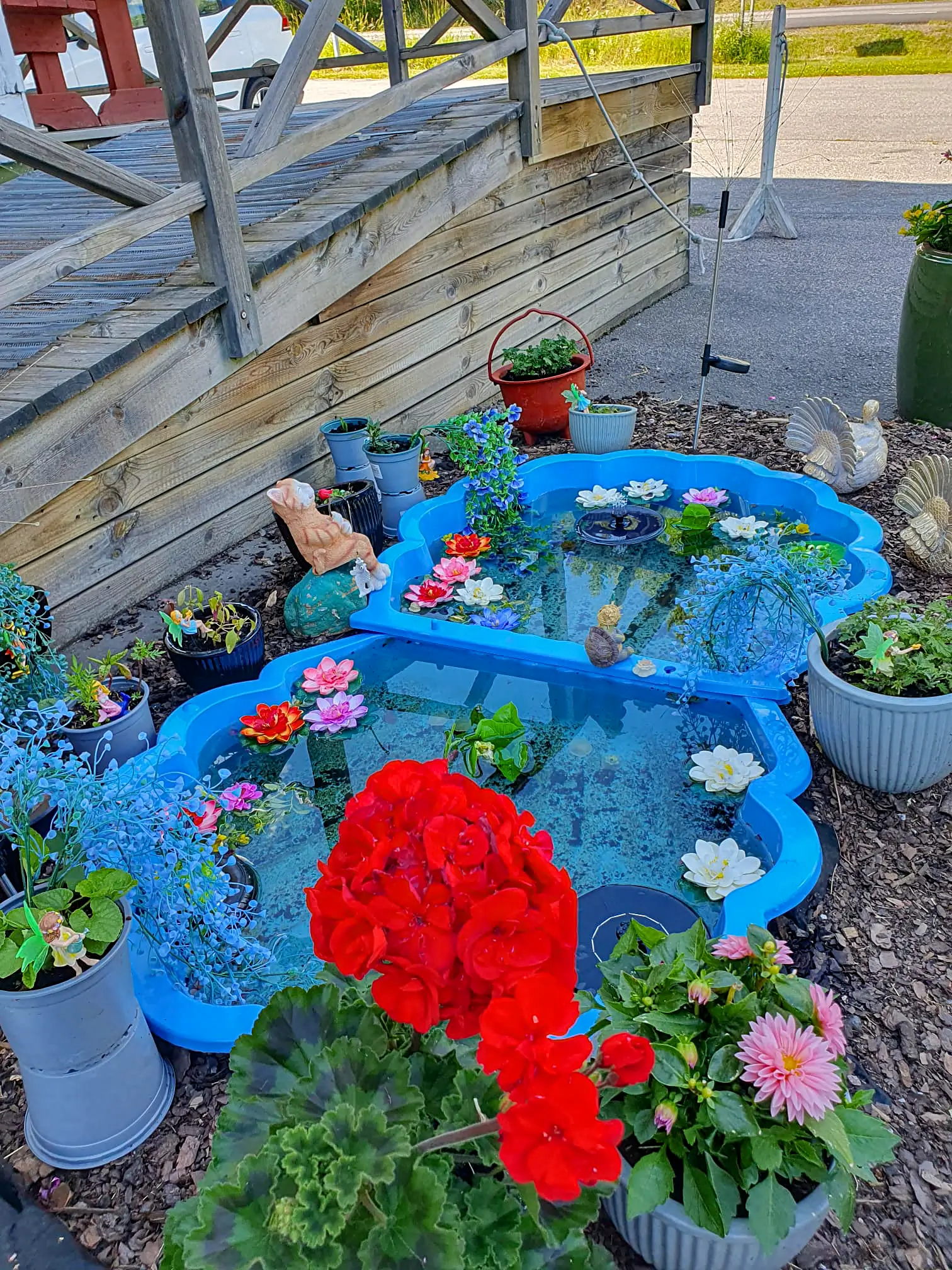 A colorful garden feature at Motel E4, showcasing a bright blue decorative pond with artificial water lilies, surrounded by vibrant red geraniums and other potted flowers.