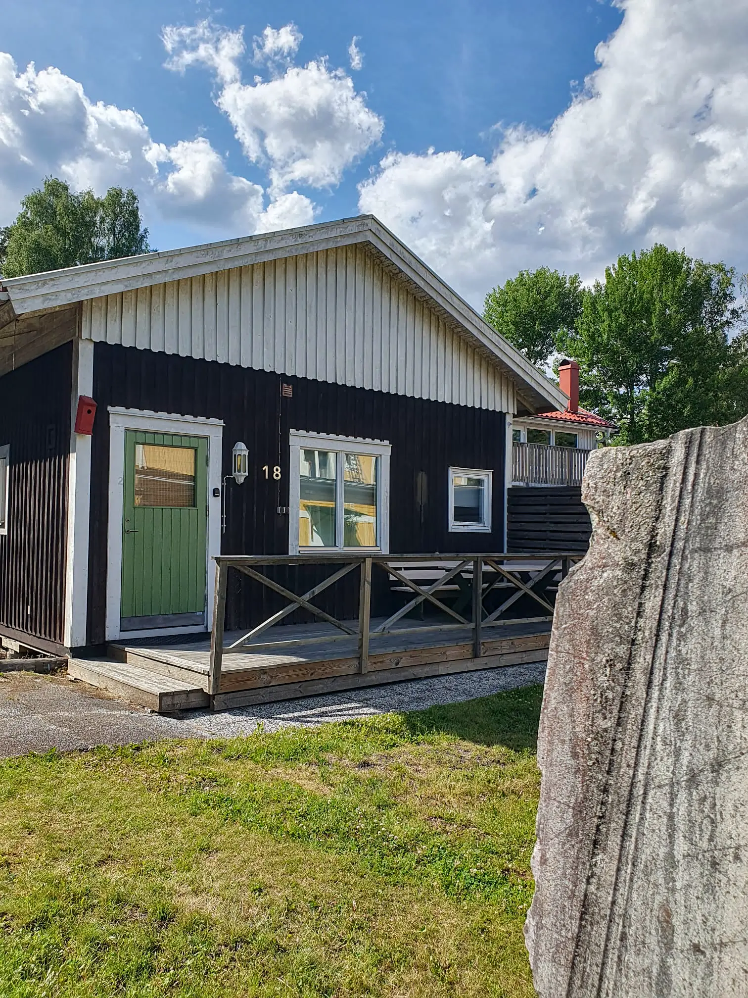 A cozy dark brown wooden cabin (unit 18) at Motel E4 featuring a light green door, a small wooden porch, and a traditional stone monument in the foreground.