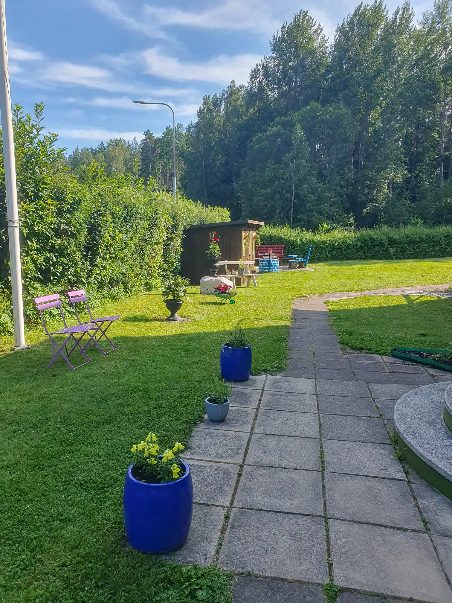 A tranquil garden area at Motel E4 featuring a stone path, blue flower pots, purple seating, and a lush forest background.